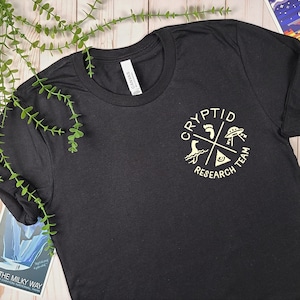 May include: Black t-shirt with a white graphic of a circle with the words "Cryptid Research Team" and four symbols: a footprint, a UFO, a sea monster, and a triangle with an eye inside.