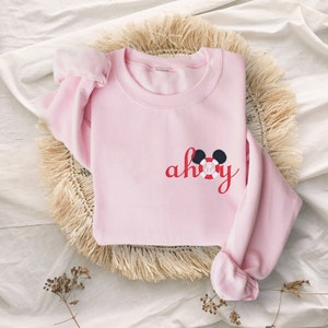 May include: A pink sweatshirt with a red "ahoy" embroidered on the front, featuring a Mickey Mouse head with a life preserver and anchor.
