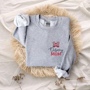 Peut inclure: Un sweat-shirt gris chin&eacute; avec un n&oelig;ud &agrave; pois rouges et blancs et l'inscription "Disney MOM" en &eacute;criture noire et en lettres rouges. Le sweat-shirt est pr&eacute;sent&eacute; sur un tapis tiss&eacute;.