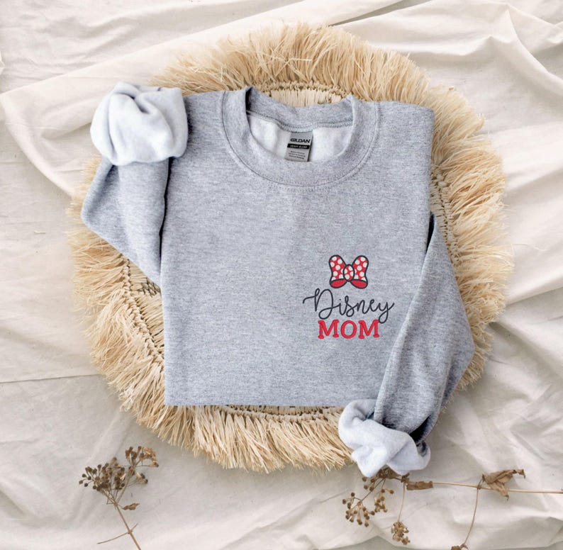 Peut inclure: Un sweat-shirt gris chin&eacute; avec un n&oelig;ud &agrave; pois rouges et blancs et l'inscription "Disney MOM" en &eacute;criture noire et en lettres rouges. Le sweat-shirt est pr&eacute;sent&eacute; sur un tapis tiss&eacute;.
