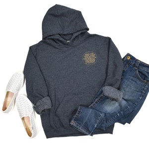 May include: A dark gray hooded sweatshirt with a gold circle graphic that says "Where you land is destiny, the world is full of unexpected turns & fairy tales". The sweatshirt is paired with blue jeans and white slip-on shoes.