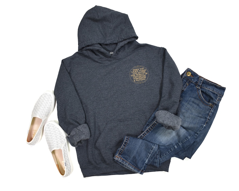 May include: A dark gray hooded sweatshirt with a gold circle graphic that says "Where you land is destiny, the world is full of unexpected turns & fairy tales". The sweatshirt is paired with blue jeans and white slip-on shoes.