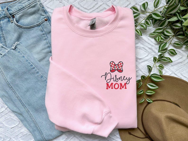 Peut inclure: Un sweat-shirt rose clair avec l'inscription "Disney MOM" en &eacute;criture noire et rouge. Un n&oelig;ud &agrave; pois rouges et blancs est plac&eacute; au-dessus du texte. Le sweat-shirt est &agrave; c&ocirc;t&eacute; d'un jean bleu et d'un chapeau marron.