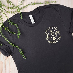 May include: Black t-shirt with a gold graphic of a circle with the text "Cryptid Research Team" and four symbols: a footprint, a UFO, a sea monster, and a triangle with an eye.