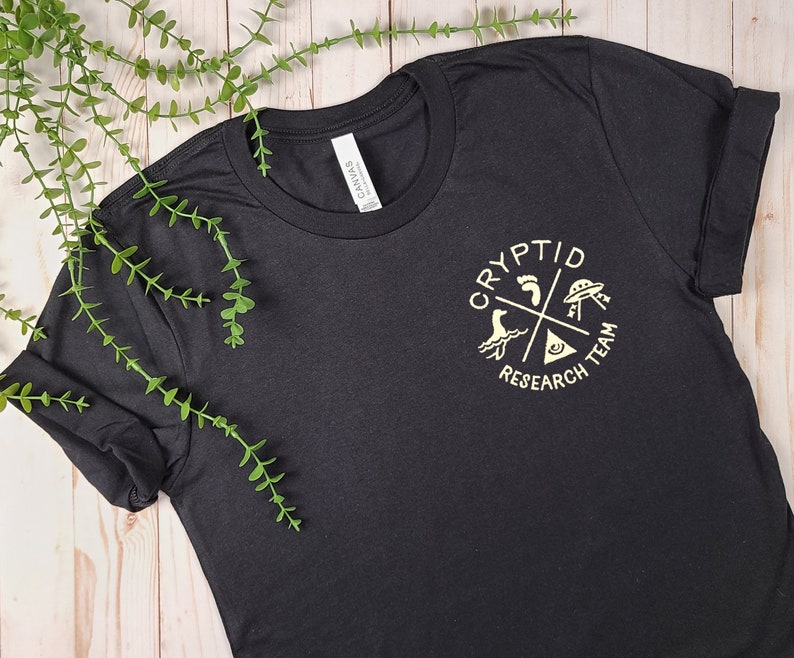 May include: Black t-shirt with a gold graphic of a circle with the text "Cryptid Research Team" and four symbols: a footprint, a UFO, a sea monster, and a triangle with an eye.