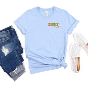 Pu&ograve; includere: Maglietta blu chiaro con il testo "DISNEY adult" in lettere colorate sul davanti. La maglietta &egrave; abbinata a jeans blu e scarpe bianche da infilare.