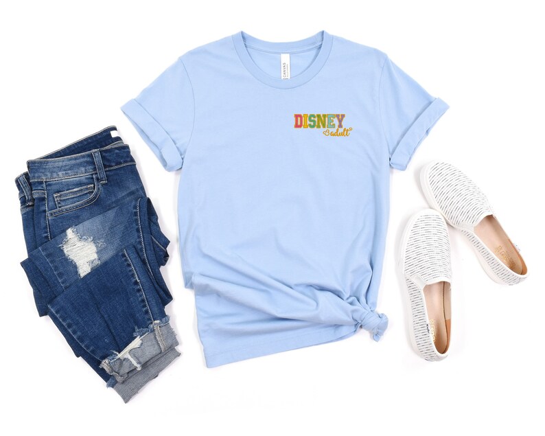 Pu&ograve; includere: Maglietta blu chiaro con il testo "DISNEY adult" in lettere colorate sul davanti. La maglietta &egrave; abbinata a jeans blu e scarpe bianche da infilare.