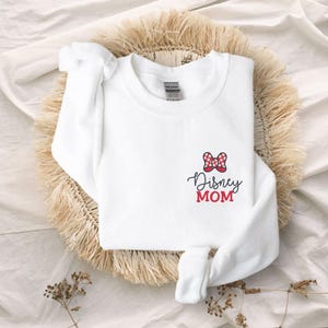 Peut inclure: Sweat-shirt blanc avec un n&oelig;ud &agrave; pois rouges et blancs et l'inscription "Disney MOM" en &eacute;criture noire et en lettres rouges. Le sweat-shirt est pli&eacute; sur un tapis tiss&eacute;.