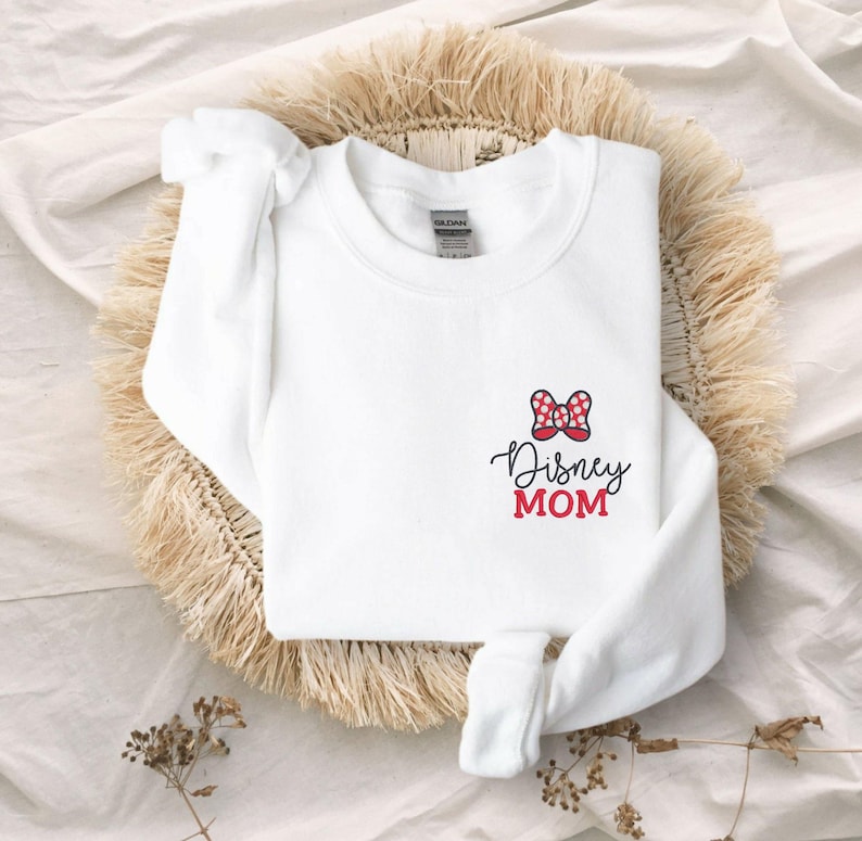 Peut inclure: Sweat-shirt blanc avec un n&oelig;ud &agrave; pois rouges et blancs et l'inscription "Disney MOM" en &eacute;criture noire et en lettres rouges. Le sweat-shirt est pli&eacute; sur un tapis tiss&eacute;.