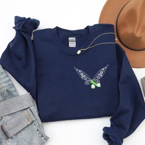 Puede incluir: Una sudadera azul marino con un diseño de mariposa bordada en blanco y verde en la parte delantera. Un collar dorado está drapeado sobre la sudadera. La sudadera se combina con unos vaqueros azules y un sombrero marrón.