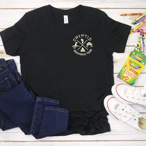 May include: Black t-shirt featuring a gold graphic of a circle with a cross inside. The circle displays the words "Cryptid Research Team" around it. The cross has a UFO, a monster, and a bird on it. The shirt is laid out on a white wooden surface with a pair of white trainers, blue jeans, and a box of crayons.