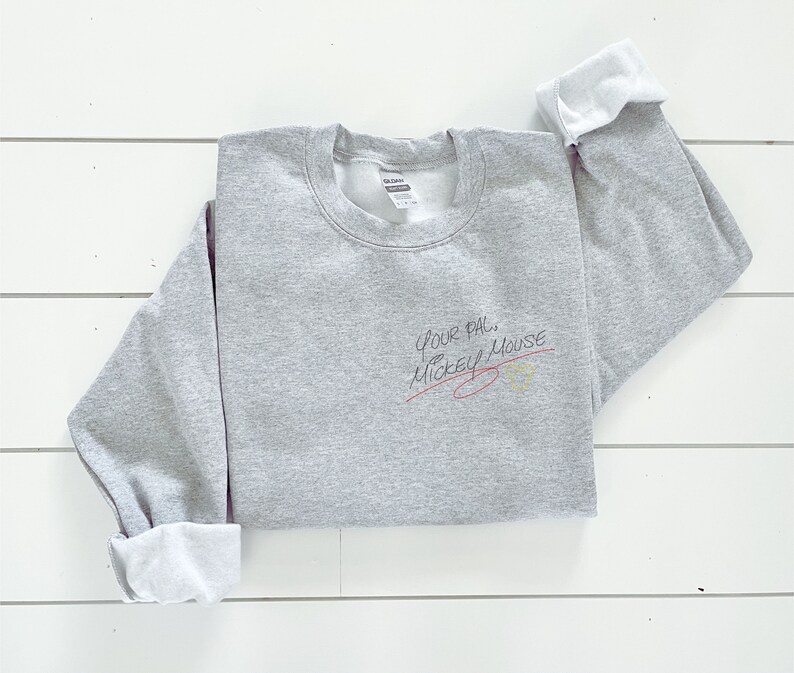 Puede incluir: Sudadera gris con el texto "Your pal, Mickey Mouse" escrito en tinta negra con una l&iacute;nea roja trazada a trav&eacute;s de &eacute;l. Un c&iacute;rculo amarillo con un contorno negro est&aacute; dibujado al lado del texto.