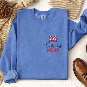 Peut inclure: Un sweat-shirt bleu avec un n&oelig;ud rouge et blanc et l'inscription "Disney MOM" brod&eacute;e sur le devant. L'image pr&eacute;sente &eacute;galement des bottes en cuir marron et un bonnet beige.