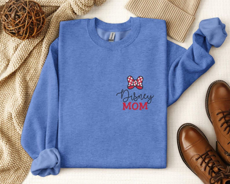 Peut inclure: Un sweat-shirt bleu avec un n&oelig;ud rouge et blanc et l'inscription "Disney MOM" brod&eacute;e sur le devant. L'image pr&eacute;sente &eacute;galement des bottes en cuir marron et un bonnet beige.
