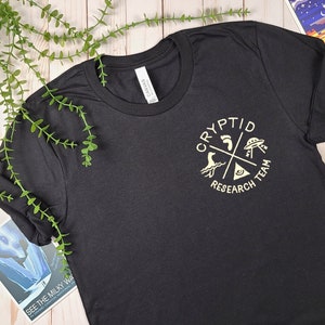 May include: Black t-shirt with a gold graphic of a circle with the words "Cryptid Research Team" and images of a footprint, a UFO, a sea monster, and a triangle with an eye inside.