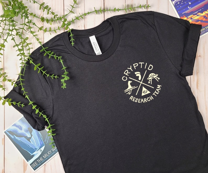 May include: Black t-shirt with a gold graphic of a circle with the words "Cryptid Research Team" and images of a footprint, a UFO, a sea monster, and a triangle with an eye inside.
