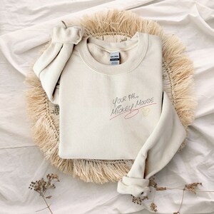 Puede incluir: Una sudadera de color crema con el texto "Your pal, Mickey Mouse" escrito en tinta negra con una l&iacute;nea roja debajo. Una cabeza de Mickey Mouse amarilla est&aacute; dibujada al lado del texto.