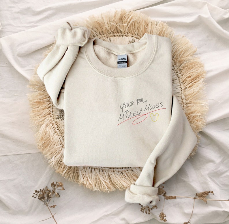 Puede incluir: Una sudadera de color crema con el texto "Your pal, Mickey Mouse" escrito en tinta negra con una l&iacute;nea roja debajo. Una cabeza de Mickey Mouse amarilla est&aacute; dibujada al lado del texto.