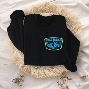 May include: Black long-sleeved sweatshirt with a blue and green embroidered patch that says "SPACE RANGERS" with a spaceship design.