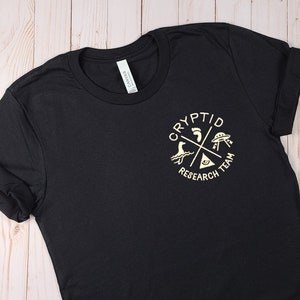 May include: Black t-shirt with a gold graphic of a circle with the text "Cryptid Research Team" and four symbols: a footprint, a UFO, a sea monster, and an eye.