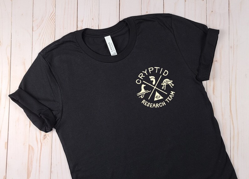 May include: Black t-shirt with a gold graphic of a circle with the text "Cryptid Research Team" and four symbols: a footprint, a UFO, a sea monster, and an eye.