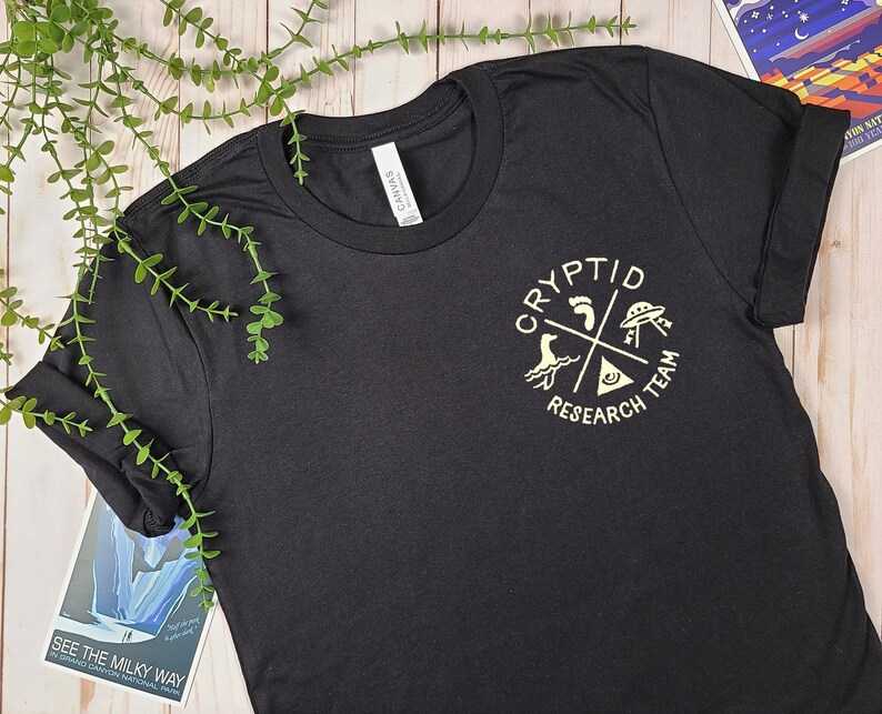 May include: Black t-shirt with a white graphic of a circle with the words "Cryptid Research Team" and four symbols: a footprint, a UFO, a sea monster, and a triangle with an eye inside.