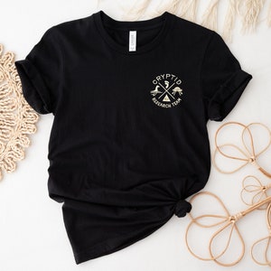 May include: Black t-shirt with a white graphic of a cross with a UFO, a monster, a ghost, and a triangle. The text "Cryptid Research Team" is written around the cross.
