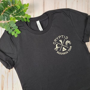 May include: Black t-shirt with a white graphic of a circle with the words "Cryptid Research Team" and four symbols: a footprint, a UFO, a sea monster, and an eye.