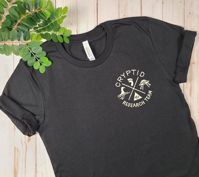 May include: Black t-shirt with a white graphic of a circle with the words "Cryptid Research Team" and four symbols: a footprint, a UFO, a sea monster, and an eye.