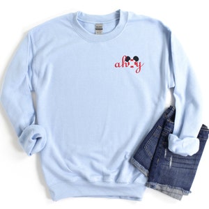 May include: Light blue sweatshirt with a red "ah-ha" text and Mickey Mouse ears on the front.