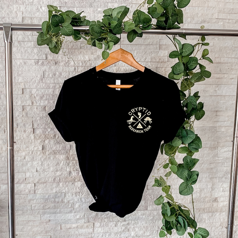 May include: Black t-shirt with a gold graphic of a cross with a UFO, a turtle, and a bird. The text "Cryptid Research Team" is written around the cross.