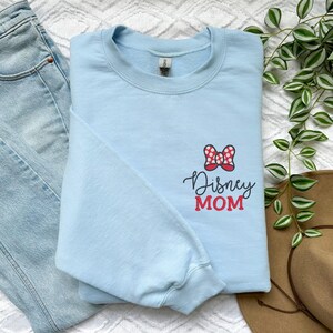 Peut inclure: Un sweat-shirt bleu clair avec un n&oelig;ud &agrave; pois rouges et blancs et l'inscription "Disney MOM". Le sweat-shirt est pli&eacute; sur un jean bleu clair et un chapeau marron.
