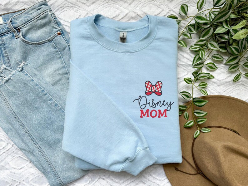 Peut inclure: Un sweat-shirt bleu clair avec un n&oelig;ud &agrave; pois rouges et blancs et l'inscription "Disney MOM". Le sweat-shirt est pli&eacute; sur un jean bleu clair et un chapeau marron.