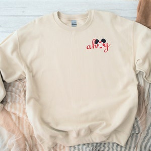 May include: A beige sweatshirt with a red embroidered design that says "ah-hy" with a Mickey Mouse head above the text.