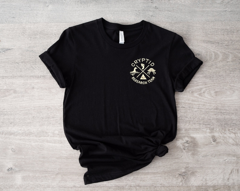 May include: Black t-shirt with a white graphic of a cross with a footprint, UFO, and a monster. The text "Cryptid Research Team" is written around the cross.