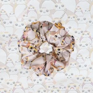 May include: A white satin scrunchie with a repeating pattern of cartoon characters. The characters are brown, white, and yellow and have large eyes.
