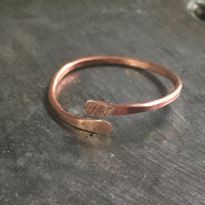 May include: A simple, minimalist copper ring with a slightly hammered finish. The ring is open and has a single, thin band that wraps around the finger.