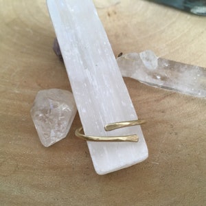 May include: A simple gold hammered metal ring with a slightly curved design. The ring is displayed on a white crystal.