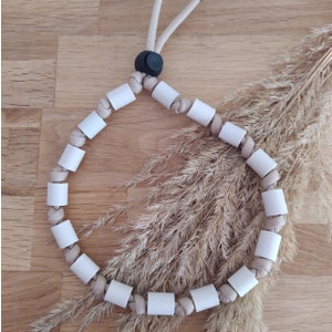 Puede incluir: Un collar de cuerda beige con cuentas de madera blancas. El collar está sobre una superficie de madera con una planta de hierba seca en el fondo.