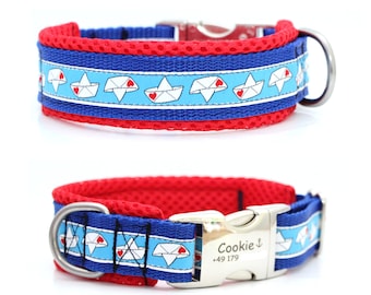 May include: A blue and red dog collar with a white and blue patterned design featuring paper boats with red hearts. The collar has a metal buckle and a D-ring for leash attachment. The collar has the name "Cookie" and a phone number.