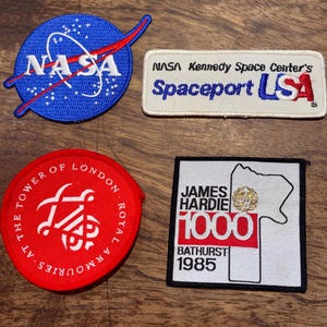 May include: Four embroidered patches. One is a blue circle with the NASA logo. Another is a white rectangle with "NASA Kennedy Space Center's Spaceport USA". A red circular patch reads "The Tower of London". A square patch reads "James Hardie 1000 Bathurst 1985".