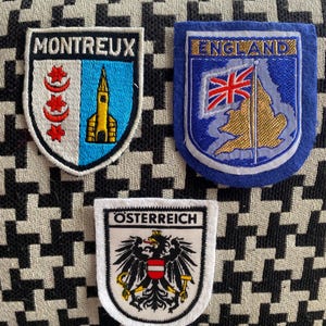 May include: Three embroidered patches. One reads "Montreux" with a church and red stars. Another says "England" with a map and flag. The third says "Österreich" with an eagle. Patches are on a patterned background.