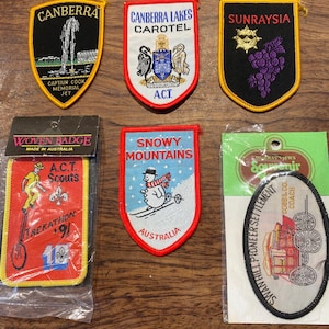 May include: Six embroidered souvenir patches featuring Australian themes. Top row: Canberra Captain Cook Memorial Jet, Canberra Lakes Carotel, and Sunraysia grapes. Bottom row: A.C.T. Scouts Trekathon 91, Snowy Mountains skier, and Swan Hill Pioneer Settlement.