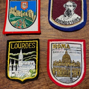 May include: Four embroidered souvenir patches featuring regional landmarks. Top left: Dover with a castle. Top right: Bleak House Broadstairs with a portrait. Bottom left: Lourdes with a cathedral. Bottom right: Roma Vaticano Basilica San Pietro.