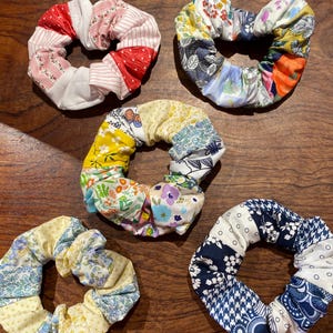 May include: Five handmade scrunchies in various patchwork patterns. The scrunchies feature fabrics in red, white, yellow, blue, and floral prints. The scrunchies are arranged on a wooden surface.