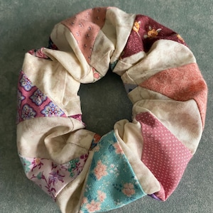 May include: A patchwork scrunchie in a variety of fabrics and patterns. The design includes floral, polka dot, and geometric prints in pink, cream, and teal. This hair accessory is a stylish addition to any outfit.