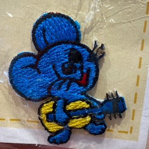 May include: Embroidered patch of a blue cartoon mouse playing a yellow and blue guitar. The mouse has black eyes and a red tongue. The patch is on a light yellow background.