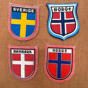 May include: Four embroidered patches displaying national flags. The top left patch reads "SVERIGE" with a blue and yellow cross. The top right patch says "NORGE" with a red and white cross. The bottom left patch says "DANMARK" with a white cross. The bottom right patch says "NORGE" with a blue and white cross.
