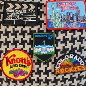 May include: A selection of embroidered patches with diverse designs. Includes a Universal Studios clapperboard, Liberty Island with the Statue of Liberty, Washington D.C., Knott's Berry Farm, and Colorado Rockies.