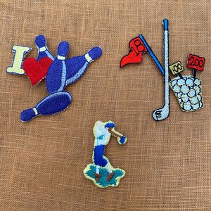 May include: Embroidered patches with sports themes. Designs include a bowling pin with a heart and the letter "I", a golf scene with a flag and yardage markers, and a baseball player swinging a bat. The patches are in blue, red, and white.